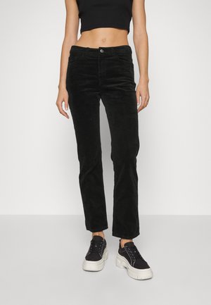 Pantalons en velours côtelé noirs avec une coupe ajustée, taille haute et détails de couture visibles. Associés à des baskets noires avec une semelle épaisse.