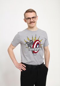 Grå bomulds t-shirt med et grafisk motiv af en hammer og et skjold med ordet "Worthy," omgivet af gule lynaccenter. Sorte bukser.