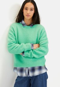 Miętowozielony, oversize'owy sweter z prążkowanymi mankietami, noszony na niebieskiej koszuli w kratę. Sweter ma miękką, teksturowaną fakturę i luźny krój.