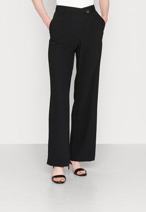 Personne portant un pantalon noir large taille haute et des talons ouverts noirs, les mains dans les poches, debout sur un sol gris clair.