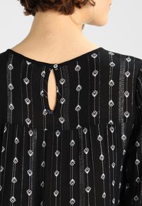 Schwarzes Kleid mit einem Schlüsselloch-Ausschnitt am Rücken, verziert mit weißer Blumenstickerei und vertikalen Streifen. Weiche Stofftextur.