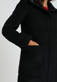 Svart ullblandad kappa med hög krage, långa ärmar och sidofickor; har en strukturerad yta och stickad detaljering.