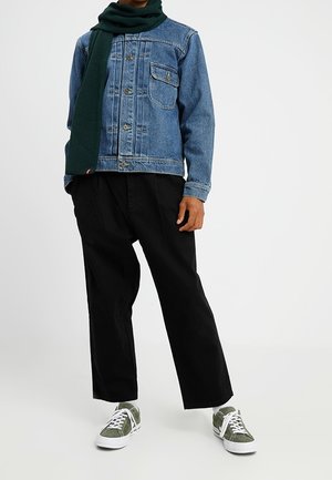 Personne portant une veste en denim bleu, un pantalon large noir, une écharpe verte et des baskets vertes à lacets blancs, debout devant un fond neutre.