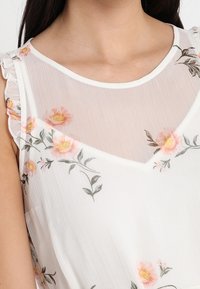 Weiße Bluse mit einem durchsichtigen, blumigen Überzug mit rosafarbenen und gelben Blumen. Rüschen an den Schultern und im V-Ausschnitt-Design. Leichter Stoff.