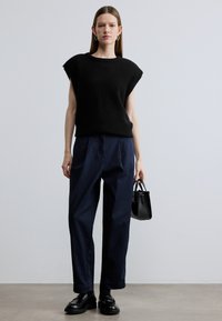 Zwarte gebreide mouwloze top met een ronde hals, gecombineerd met losse marineblauwe broek. Accessoires zijn onder andere een kleine zwarte handtas en loafers.