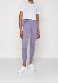 Kobieta w białej, krótkiej siateczkowej koszulce, fioletowych legginsach z wysokim stanem i bocznymi kieszeniami, pasującej torbie na biodra oraz białych sneakersach, stoi.