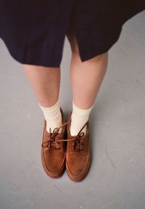 Scarpe in suede marrone con lacci e suola piatta, indossate con calzini giallo chiaro a quadri. In piedi su un pavimento grigio.