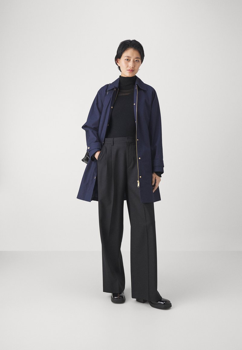 Abrigo azul marino con detalles dorados, superpuesto a un cuello de tortuga negro y pantalones negros de pierna ancha; unos zapatos negros planos completan el conjunto.