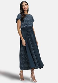 Navyblauwe maxi-jurk met korte mouwen, geplooide taille en een verticaal wit stippenpatroon. Gecombineerd met witte sandaaltjes met bandjes. Glad textuur van de stof.