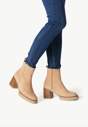 Zamszowe botki za kostkę w jasnym beżu z grubym obcasem, z bocznym zamkiem błyskawicznym i fakturowaną podeszwą dla lepszej przyczepności, noszone z ciemnoniebieskimi dżinsami.