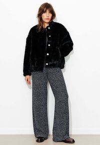 Veste en fausse fourrure noire avec un col arrondi et de gros boutons. Associée à un pantalon large bleu marine avec un motif floral blanc.