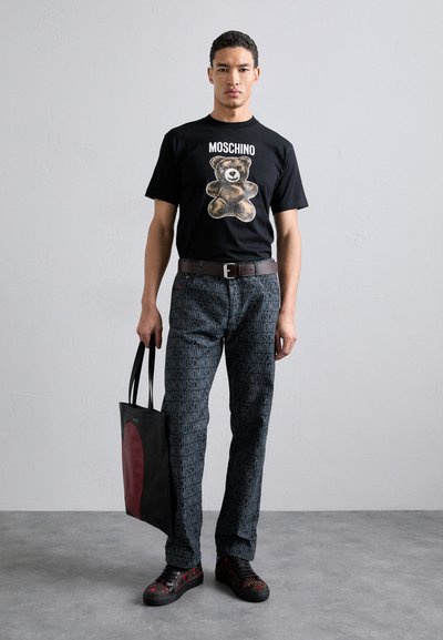 T-shirt noire avec graphique d'ours et texte "MOSCHINO", pantalon noir à motifs, sac noir avec accent rouge, baskets noires et rouges. Look décontracté dans l'ensemble.