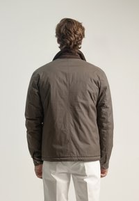 Chaqueta aislante marrón con cuello acanalado, corte ligeramente holgado y detalles de puños abotonados, combinada con pantalones blancos.