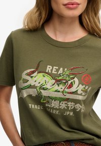 Olijfgroene katoenen t-shirt met een kleurrijke draakafbeelding, getextureerde versieringen en witte bedrukte tekst. Ronde halslijn en korte mouwen.