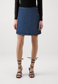 Denim mini suknja u tamnoplavoj boji s ravnim krojem, s neurednim rubom i dva dekorativna gumba sa strane. Uparena s crnim sandalama na visoku petu.