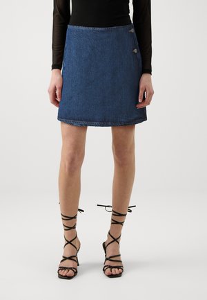 Denim mini suknja u tamnoplavoj boji s ravnim krojem, s neurednim rubom i dva dekorativna gumba sa strane. Uparena s crnim sandalama na visoku petu.