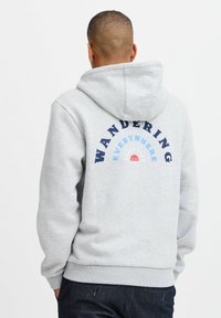 Grå hettegenser med kengurulomme, med et trykt design på baksiden som leser "VANDRING OVERALT" i blått og rødt.