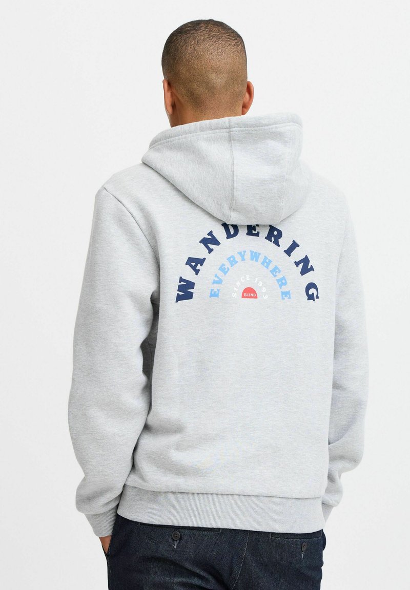 Grå hettegenser med kengurulomme, med et trykt design på baksiden som leser "VANDRING OVERALT" i blått og rødt.