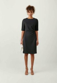 Vestido preto com mangas curtas, decote redondo, tecido texturizado, até ao joelho, usado com sapatos de salto nude e uma mala de mão branca.