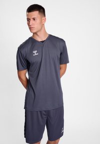 Γκρι αθλητική μπλούζα με κοντά μανίκια και λαιμόκοψη σε σχήμα V, που διαθέτει λογότυπο στην αριστερή πλευρά. Συνδυάζεται με γκρι σορτσάκι, και τα δύο κατασκευασμένα από λεία ύλη.