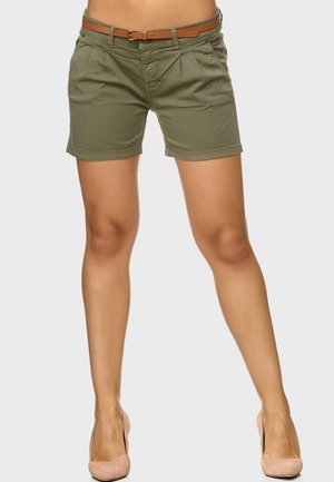 Kobieta w oliwkowych szortach z paskiem i cielistych szpilkach, stojąca na tle jasnego, gładkiego tła.