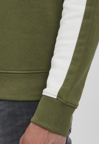 Felpa verde oliva con orlo a coste testurizzato e striscia bianca a contrasto. La ripresa ravvicinata mostra i dettagli della manica su uno sfondo di tessuto scuro.
