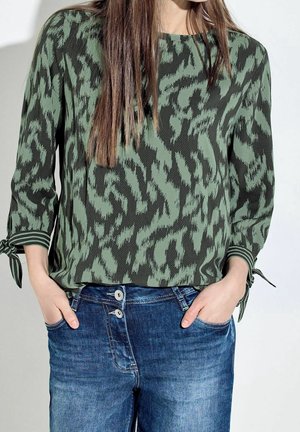 Donna che indossa una blusa a motivi verdi e neri con maniche legate e jeans blu, mani nelle tasche, su uno sfondo semplice.