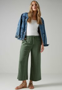 Gröna vida byxor med elastisk midja, tillsammans med en vit linne och en slitna denimjacka, samt enkla bruna sandaler.