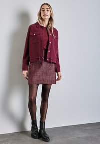 Chaqueta corta burdeos con dos bolsillos en el pecho, combinada con una minifalda de cuadros texturizada y botas negras hasta el tobillo con tacón grueso.