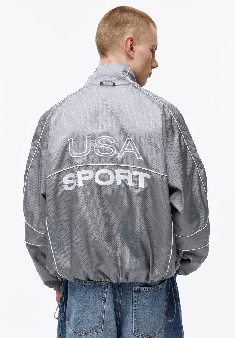 Mladý muž v lesklej šedej športovej bunde s nápisom "USA SPORT" na chrbte, v modrých džínsových nohaviciach, stojaci chrbtom.