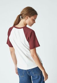 Maglietta raglan bianca e bordeaux con maniche corte, caratterizzata da un collo rotondo e una vestibilità rilassata, su uno sfondo semplice.