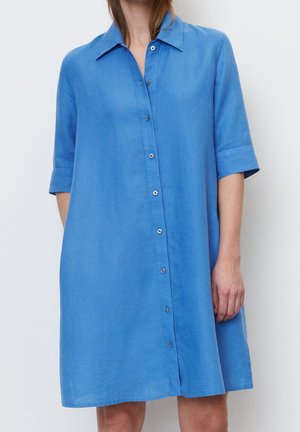 Vrouw draagt een blauw overhemdjurk met knopen, korte mouwen en een kraag, die tot op de knie reikt, staand tegen een effen witte achtergrond.