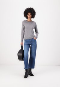 Grå långärmad tröja i kombination med vida blå jeans och svarta klackstövlar, bärande på en rund svart väska. Minimal bakgrund.