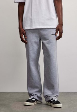Person iført grå løse joggingbukser med sorte og hvide sneakers og en hvid kortærmet skjorte, der står på et betongulv.