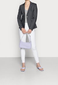 Grå skräddarsydd kavaj, grå linne, vita jeans, lila handväska med texturerat mönster och lila flip-flop, stående mot en enkel bakgrund.