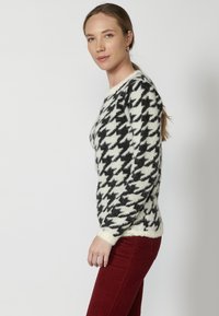 Pulover cu model houndstooth alb-negru, cu o textură pufoasă, având guler rotund și manșete ribbed, combinat cu pantaloni roșii.