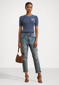 Pull en tricot bleu à manches courtes avec motif en câble et logo brodé, associé à un jean délavé avec des éclaboussures de peinture et un sac à main marron.