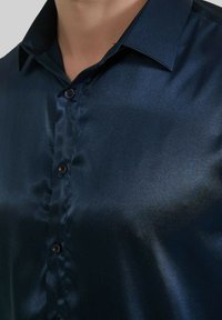 Chemise boutonnée en satin bleu marine avec une texture brillante, col classique et six boutons sur le devant. Ajustement lisse et taillé.