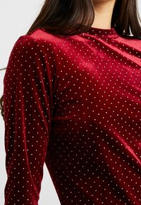 Sammetstopp i djupröd färg, med rund halsringning, långa ärmar och ett mönster av små, glänsande silverprickar över tyget.