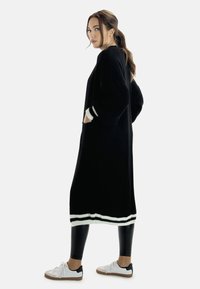 Cardigan largo negro de punto con acentos de rayas blancas, combinado con leggings negros y zapatillas blancas. El cardigan tiene un corte relajado y bolsillos.