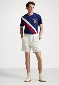 Μπλε ναυτικό μπλουζάκι με λευκή και κόκκινη διαγώνια ρίγα, λογότυπο "USA". Λευκά πλισέ σορτς. Ανοιχτόχρωμα παπούτσια με ανοιχτόχρωμες κάλτσες. Απλός σχεδιασμός.