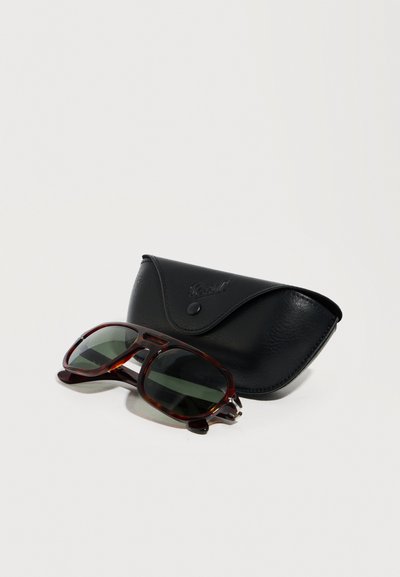 Γυαλιά ηλίου με καφέ σκελετό σε σχήμα χελώνας και σκούρους φακούς, τοποθετημένα δίπλα σε μια σκληρή θήκη από μαύρο δέρμα πάνω σε λευκή επιφάνεια.