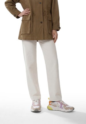 Person iført beige bukser med vide ben, pastelfarvede multifarvede sneakers og en brun jakke med store frontlommer, stående med den ene hånd på hoften.