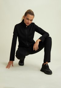 Giacca sportiva nera con colletto alto, zip e design aderente, abbinata a pantaloni neri a sigaretta e sneaker neri chunky.