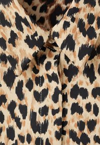 Límec košile s leopardím vzorem v hnědé, černé a hnědé nepravidelné skvrny a viditelným malým knoflíkem na výstřihu.