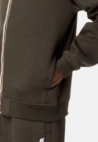 Felpa zip con cappuccio verde oliva, con polsini e orlo a costine, caratterizzata da una zip multicolore e tasche laterali. Tessuto morbido al tatto.