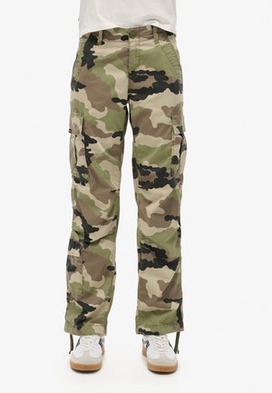 Camo cargobroek in groen en beige met zwarte accenten, voorzien van zijzakken, een relaxte pasvorm en verstelbare manchetten. Gecombineerd met witte sneakers.