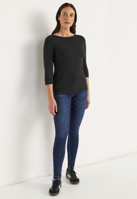 Schwarzes, figurbetontes Longsleeve-Oberteil mit Boot-Ausschnitt, kombiniert mit dunkelblauen Skinny-Jeans und schwarzen Sportschuhen mit weißen Akzenten.