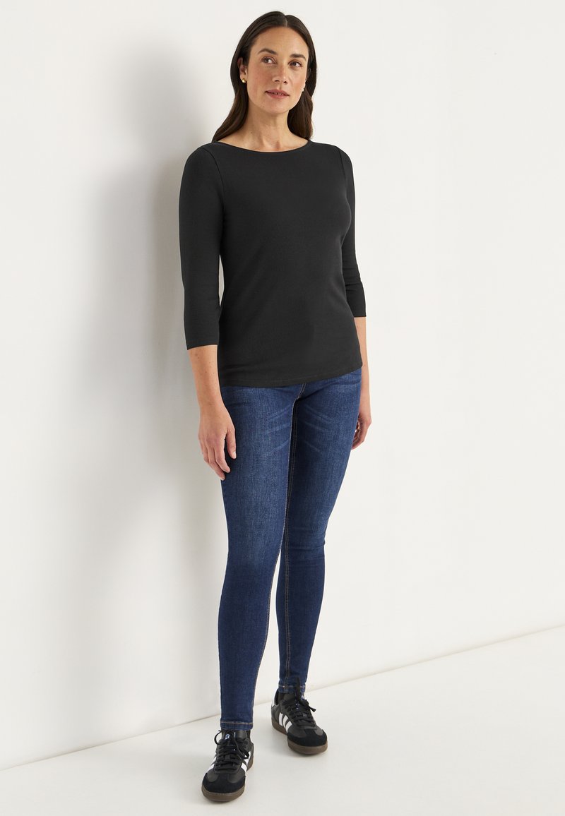 Schwarzes, figurbetontes Longsleeve-Oberteil mit Boot-Ausschnitt, kombiniert mit dunkelblauen Skinny-Jeans und schwarzen Sportschuhen mit weißen Akzenten.