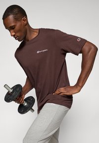 Mand i brun Champion shirt og grå sweatpants løfter en sort håndvægt, poserer med den ene hånd på hoften foran en ensfarvet baggrund.